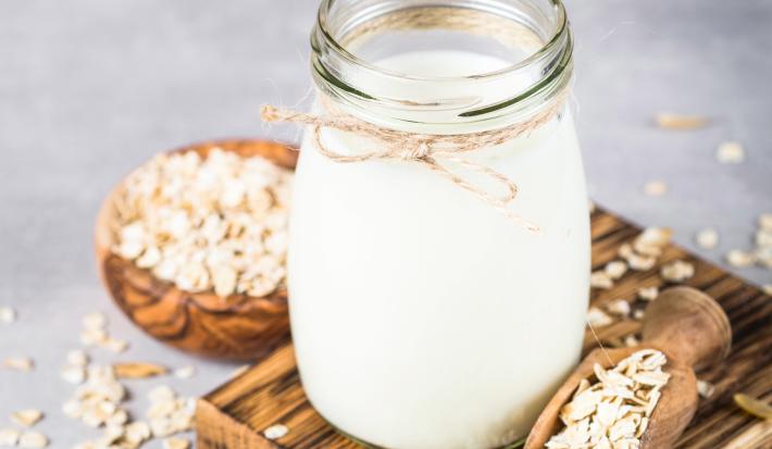 Fotografia em tons claros de um pote decorativo com bebida vegetal de coco sobre uma mini tábua de madeira escura retangular. Ao lado do pote tem aveia em flocos espalhadas e dentro de uma colher de madeira longa.