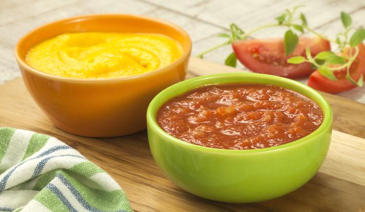 Fotografia em tons de verde, amarelo e vermelho com dois potes ao centro, sendo um amarelo com creme amarelo e um verde com um creme de tomate, ambos sobre peça de madeira, ao lado guardanapo e fatias de tomate, tudo sobre bancada de madeira.