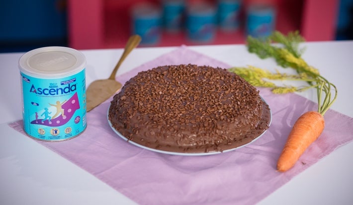 Fotografia de um bolo de cenoura com cobertura de chocolate e ascenda sobre um prato raso grande. Ao redor tem uma cenoura inteira, uma espátula e a alta de ascenda, sobre um pano roxo quadrado.