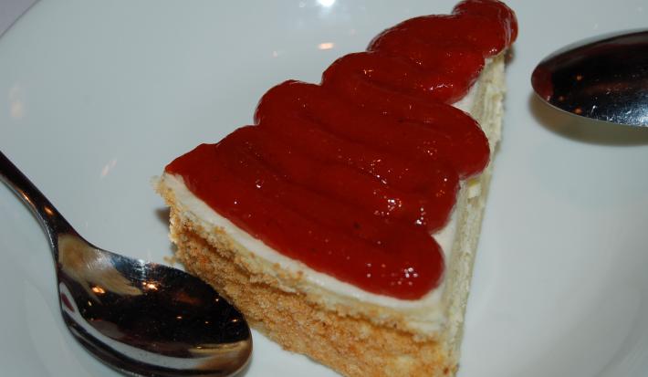 Fotografia de uma fatia de torta de ricota com leite MOÇA e cobertura de goiabada em um prato de vidro branco. Ao redor da sobremesa e apoiadas no prato tem duas colheres de sobremesa.