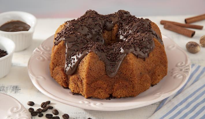 Foto da receita de Bolo de Cenoura com Especiarias Coberto com Brigadeiro de Café. Observa-se um bolo inteiro em cima de um prato de porcelana branco com brigadeiro por cima e granulado. Grãos de café, canela em pau e o brigadeiro decoram a foto.