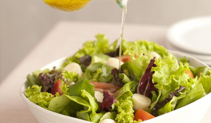 Fotografia em tons de verde e branco de uma bancada de madeira branca com um  recipiente branco quadrado com salada de folhas e um fio de azeite sendo despejado. Ao lado uma tabua de madeira com uma colher de servir.