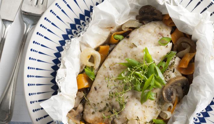 Fotografia em tons de azul em uma mesa de madeira azul com um prato branco fundo com detalhes azuis e o filé de peixe com legumes dentro dele.