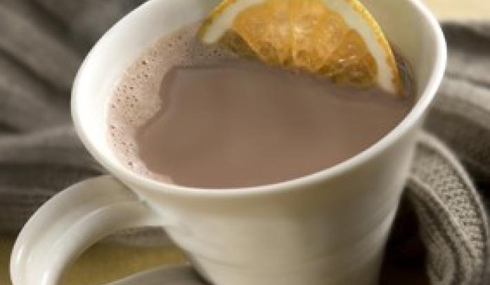 Fotografia em tons de cinza em uma bancada de madeira clara, um pano cinza escuro, uma xícara branca com o chocolate quente com uma rodela de laranja dentro.
