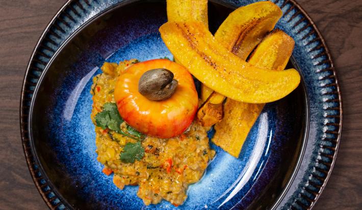 Fotografia em tons de marrom com um prato preto ao centro. Em cima do prato existe uma receita de ceviche de caju com castanhas e chips de banana da terra