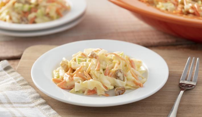 Imagem de um prato branco com a salada coleslaw de repolho. Ao lado um garfo e ao fundo mais um prato e uma travessa laranja.