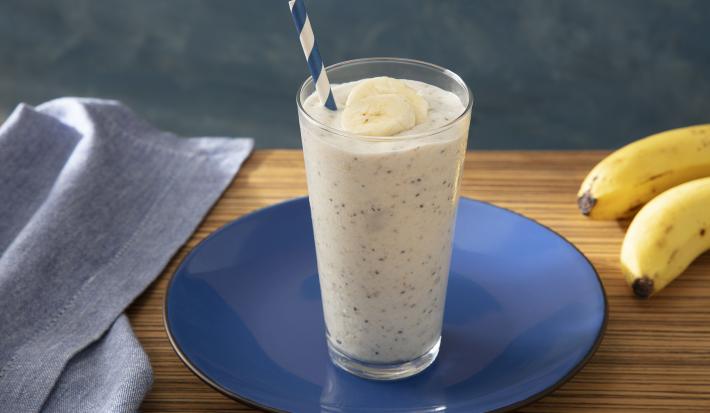 Foto da receita de Smoothie de Banana com Farinha Láctea Nestlé. Observa-se um copo com a bebida e um canudo azul e branco. Ao lado direito, um cacho de bananas