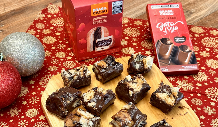 Foto da receita de palha italiana de mini panettone brasil cacau, servida em pedaços pequenos sobre uma tábua de madeira, em uma bancada decorada com itens natalinos e produtos da marca brasil cacau ao redor