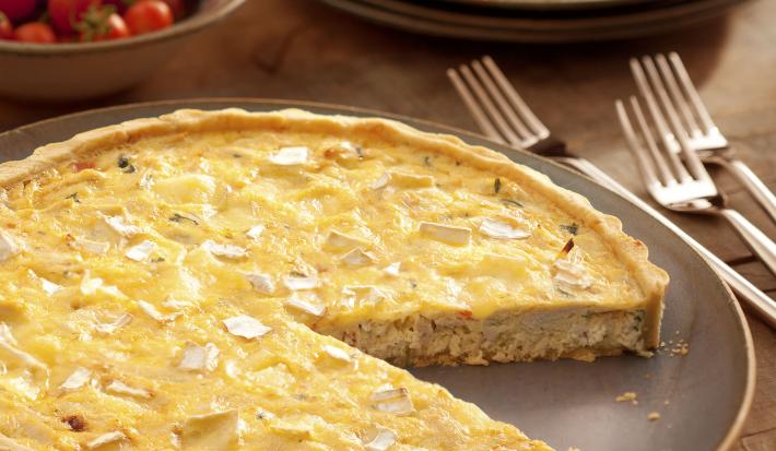 Fotografia em tons de marrom, terrosos e vermelho, com prato em tom terroso contendo uma quiche fatiada e em seu entorno três garfos, pratos empilhados com um pedaço de quiche, potinho com tomate-cereja, tudo sobre bancada de madeira.