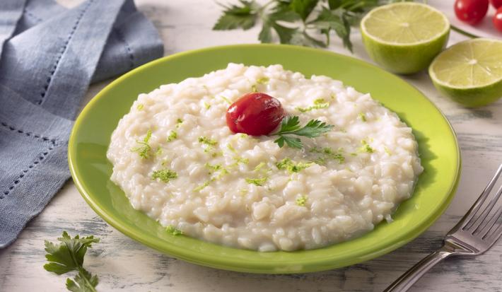 Fotografia em tons de verde em uma bancada de madeira de cor branca. Ao centro, um prato verde contendo o arroz. Ao lado, há um garfo e um pano azul. Ao fundo, há um limão partido ao meio, tomate cereja e salsinha espalhados.