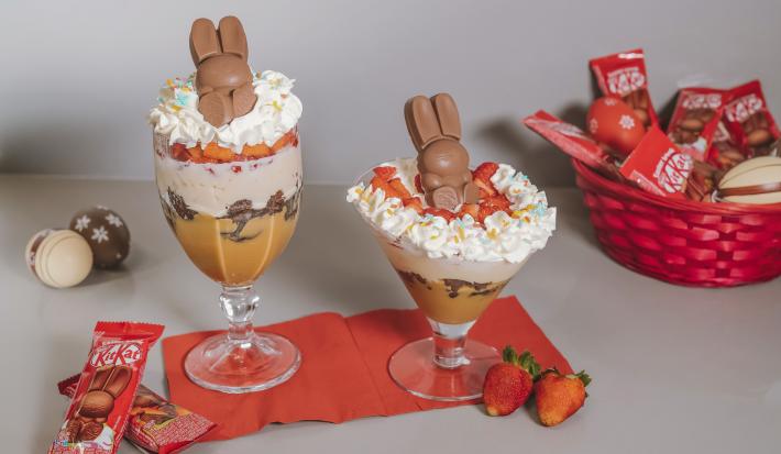 Fotografia em branco e vermelho de uma bancada branca com um paninho vermelho, sobre ele duas taças com kitkat bunny. Ao lado uma cesta com Kitkat e morangos.
