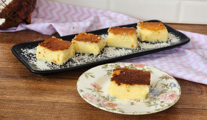 Foto da receita de Docinho do Céu Cremoso. Observa-se um recipiente preto com quatro retângulos do doce polvilhado de coco e, à frente, um pratinho de sobremesa com uma fatia também.