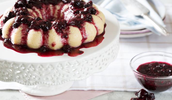 foto em tons de branco e vinho tirada de uma mesa branca, contém um suporte para servir sobremesas branco com o Brigadeirão Branco com Calda de Frutas Vermelhas. Ao lado um pote transparente redondo com a calda de frutas vermelhas.