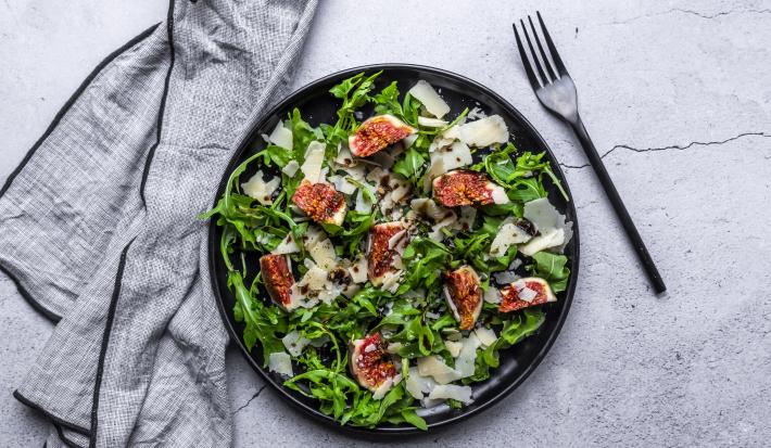 Fotografia vista de cima de uma salada de folhas, figo, requeijão e molho de maracujá e com vinagre balsâmico. A salada está em um prato raso preto, e ao lado, um garfo preto, sobre uma mesa com tons de cimento.