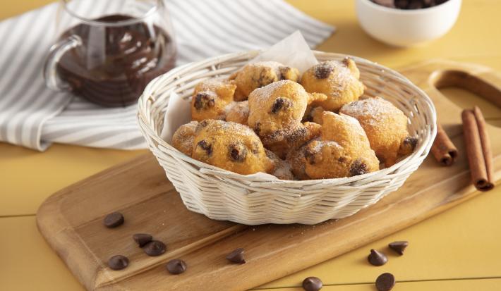 Fotografia em tons de amarelo em uma bancada de madeira com um pano listrado em cinza ao lado e um tábua de madeira no meio apoiando uma cesta de vime com bolinhos de chuva de cenoura com chocolate. Ao fundo um potinho de vidro com chocol