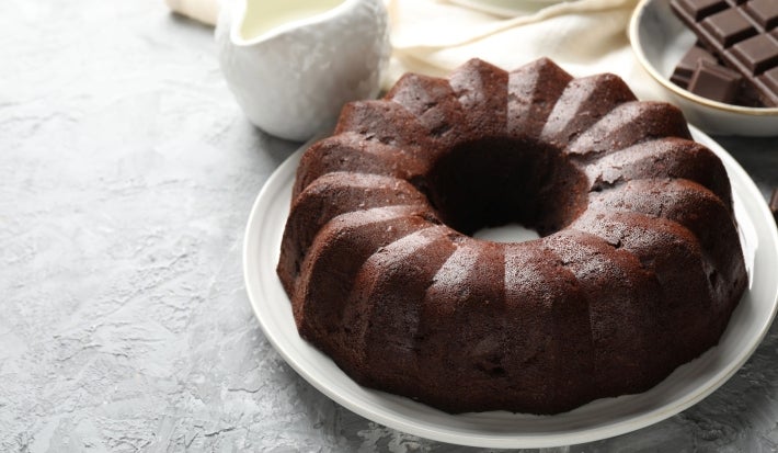 Bolo de chocolate bundt, com cor rica e textura úmida, em um prato branco. Ao fundo, há um bowl com pedaços de chocolate e um jarro de cerâmica.