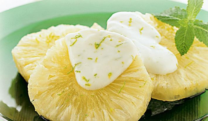 Fotografia em tons de verde em uma mesa com um pano verde, um prato redondo raso de vidro verde e três rodelas de abacaxi com o molho de iogurte com hortelã em cima dele.