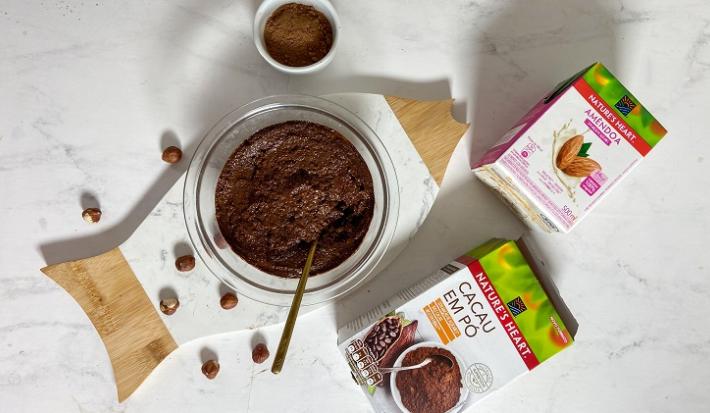 Foto em tons de branco da receita de creme de avelã caseiro servida em um bowl de vidro sobre uma tábua de madeira com embalagens de bebida vegetal de amêndoa e cacau em pó NATURE'S HEART