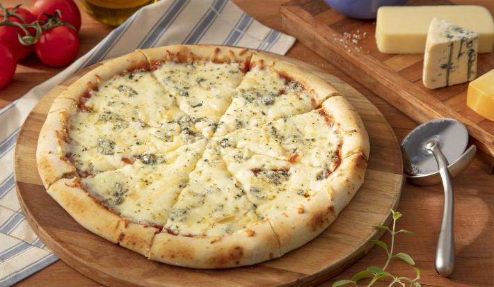 Fotografia em tons de azul em uma bancada de madeira, um pano azul listrado, uma tábua de madeira de pizza, um cortador de pizza ao lado e pedaços de queijo.