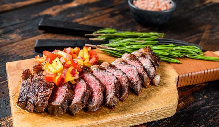 Fotografia em tons de marrom em uma bancada de madeira preta. Ao centro, uma tábua de madeira contendo a picanha fatiada ao fundo há duas facas, temperos e um recipiente contendo sal rosa.