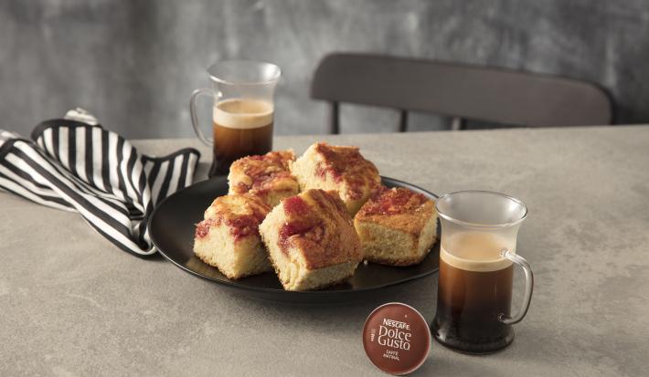 Fotografia em tons de marrom em uma bancada de madeira com um pano de prato com listra marrons, um prato redondo raso marrom com pedaços de bolo de goiabada. Ao lado, uma xícara de café Nescafé Dolce Gusto.