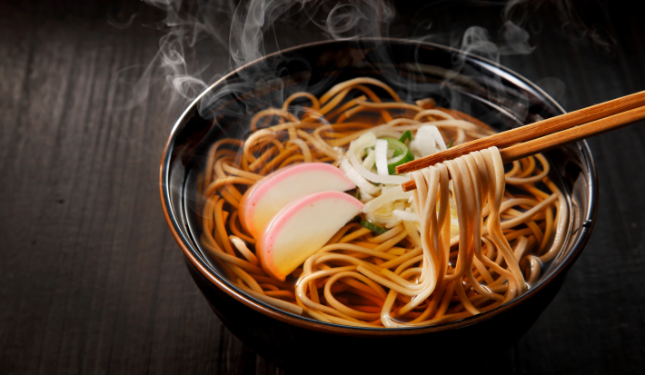 Fotografia mostra um macarrão japonês sendo segurado por hashi, ao fundo um bowl escuro com macarrão dentro.