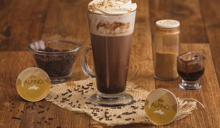 Fotografia em tons de marrom em uma bancada de madeira com um copo grande de vidro com a bebida de Alpino, chocolate quente. Ao fundo, cápsulas de alpino da Dolce Gusto, um vidrinho de canela, um copinho com raspas de chocolate.