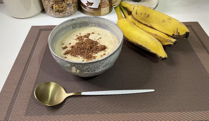 Imagem da receita de Mingau Proteico em um bowl, sobre uma mesa e ao lado uma banana e colher