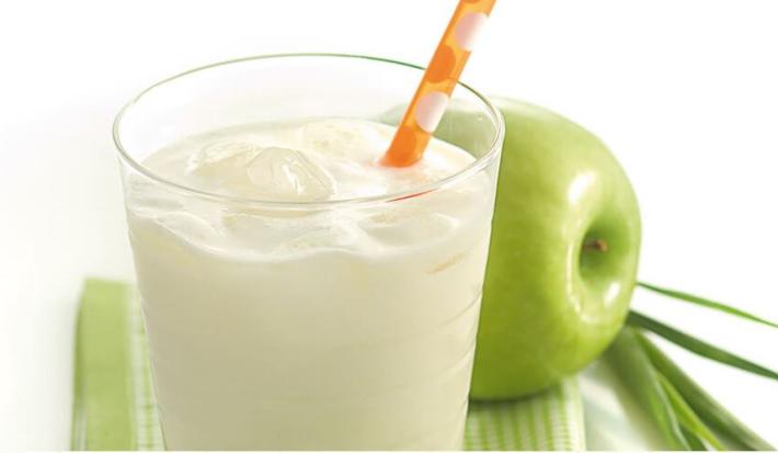 Fotografia em tons de verde e branco, ao centro como com creme branco e canudo sobre guardanapo em tons de branco e verde, ao lado maçã verde e ramo de erva-cidreira, tudo sobre bancada branca.