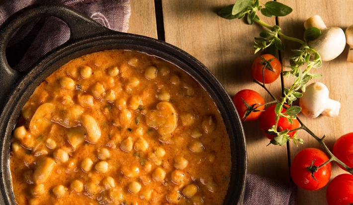 Fotografia em tons de amarelo em uma bancada de madeira com um pano azul e uma panela de ferro preta com alças laterais e o strogonoff de grão-de-bico dentro. Ao lado, tomates, champignon e salsinha espalhados na bancada.