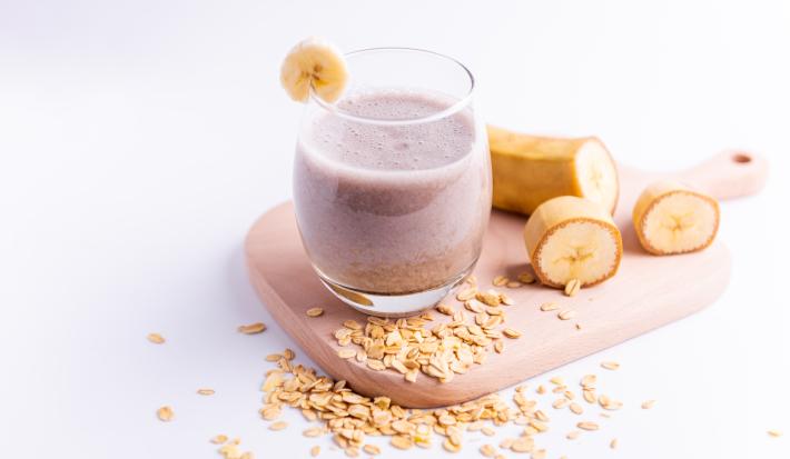 Fotografia de um copo de vidro com uma bebida de banana com leite MOÇA e neston. Na borda do copo, tem uma rodela de banana, assim como ao lado do copo.
