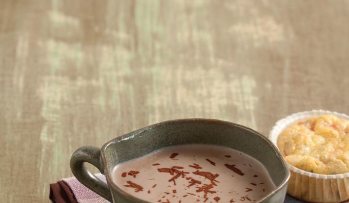 Fotografia em tons de marrom em uma bancada de madeira com um pano xadrez roxo com uma caneca grande com o chocolate quente feito com bombom de Alpino.