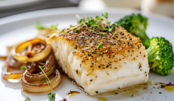 Em cima de uma mesa, prato com posta de bacalhau, cebola caramelizada  e floretes de brócolis.