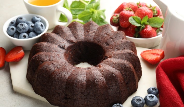 Bolo de chocolate em forma de bundt, com superfície brilhante e densa, servido em uma tábua clara. Ao redor, chá, folhas de hortelã e morangos frescos.