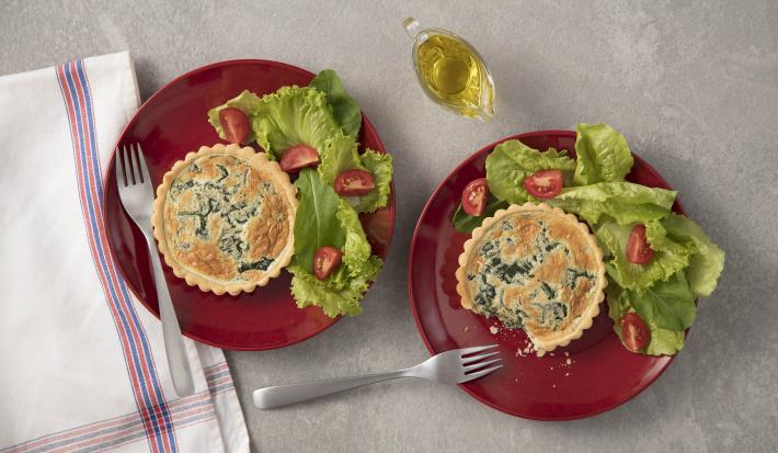 Fotografia em tons de cinza, branco, vermelho e verde de uma bancada cinza vista de cima. Contém dois pratos redondos vermelhos ambos com uma tortinha e ao lado folhas de alface com fatias de tomate e garfos para servir. Ao lado temos um pano branco.