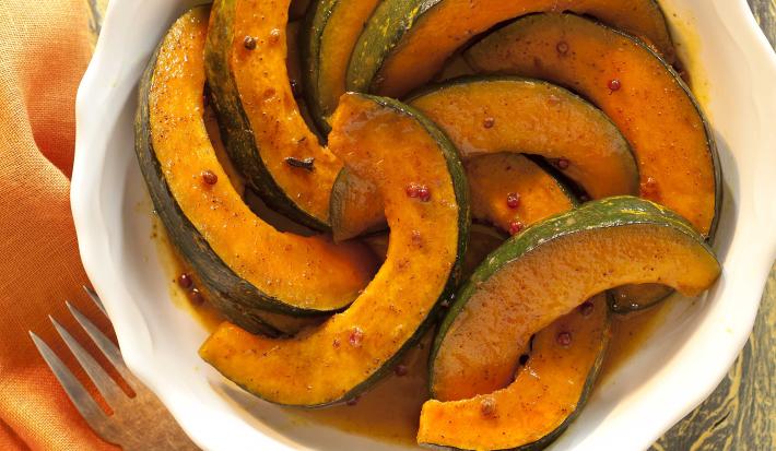 Fotografia em tons de laranja em uma bancada de madeira, ao lado um garfo apoiado em um guardanapo laranja e ao centro uma travessa redonda branca com as abóboras caramelizadas dentro.