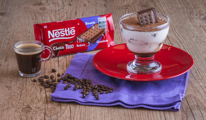 Fotografia de uma taça transparente sobre um prato redondo vermelho, preenchida com um creme branco, um pedaço de choco trio e cacau polvilhado por cima