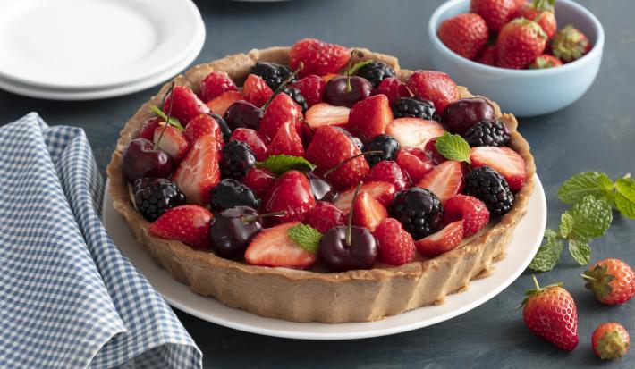 Fotografia em tons de vermelho e azul de uma bancada, ao centro a torta com morango, amoras e cerejas. Ao lado um pano quadriculado azul e branco e morangos.