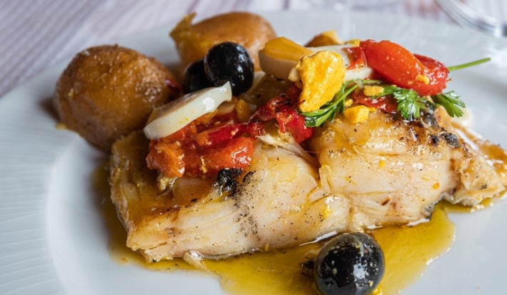 Em cima de uma mesa, prato com porção de bacalhau com azeite e tomate, pimentão, batatas e azeitonas.