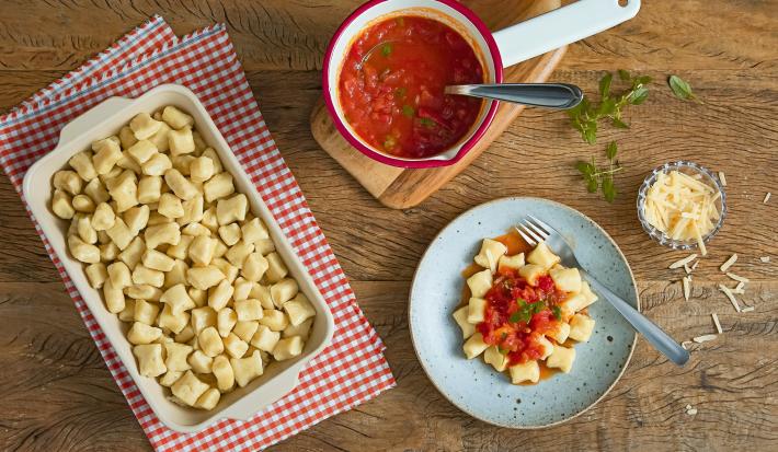 Imagem vista de cima de uma travessa com nhoques, uma panela com molho de tomate, um prato com nhoque com molho de tomate e manjericão e um potinho de queijo ralado.