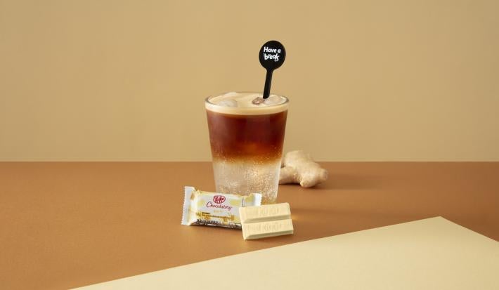 Foto da receita de Espresso Tônica. Observa-se um copo alto com gelo e duas fases aparentes: metade tônica e metade café. O cenário é bege e está decorado com gengibre e servido com um KIT KAT Branco.