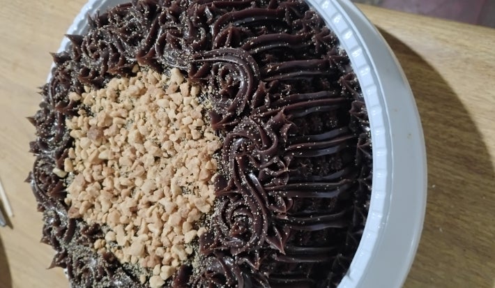 Bolo de chocolate com cobertura cremosa em rosetas e crocante granulado no centro, sobre bandeja branca.