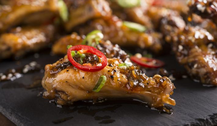 Fotografia de frango camponês dourado e brilhante com salsinha, gergelim e uma rodela de pimenta vermelha em cima, sobre um apoio preto. Ao fundo, mais unidades do frango.