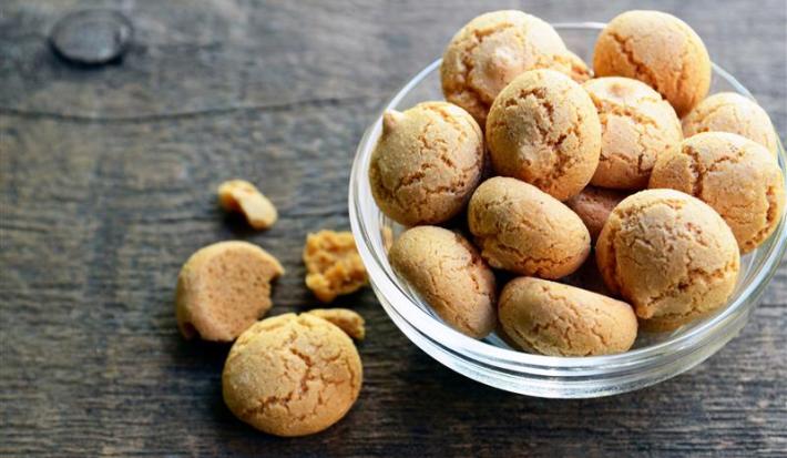 Fotografia de um recipiente de vidro médio fundo com biscoitos de amêndoas tipo amaretti, ao lado tem um e um pedaço sobre uma mesa de madeira.