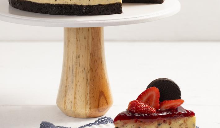 Fotografia em tons de branco em uma bancada de madeira em tons de branco com um pano marrom. No centro, um prato branco contendo uma fatia do cheesecake com um garfo ao lado e atrás está o restante do cheesecake.