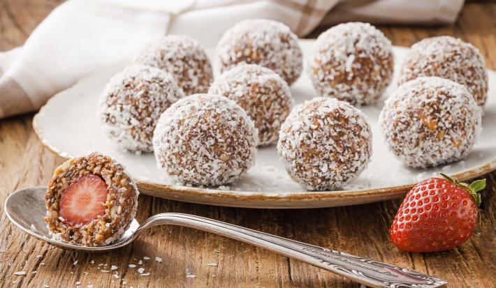 Fotografia em tons de branco, marrom e vermelho, ao centro prato branco com bombons enrolados em coco ralado, no entorno colher com bombom aberto ao meio, um morango, guardanapo branco com listras marrom, tudo sobre bancada de madeira rústica.