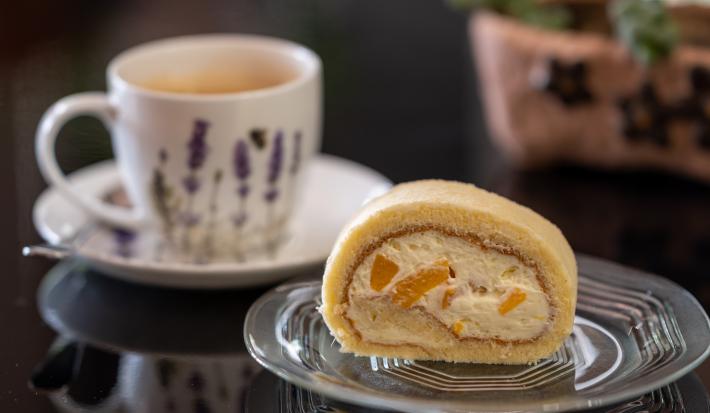 Fotografia de um rocambole feito de pão de ló de chocolate recheado com chantilly e peras cozidas, que está em um prato pequeno de vidro. Ao fundo, tem uma xícara de chá branca, sobre um miniprato de vidro branco, ao lado de uma colher de chá.