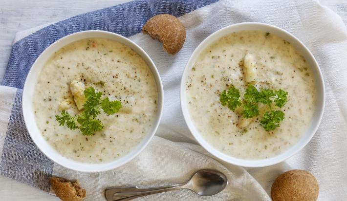 Fotografia vista de cima de dois recipientes na cor branca com um creme de aveias com palmito, pimenta por cima e salsinha. Ao redor, três biscoitos e uma colher de sopa, sobre uma toalha de mesa branca com faixas em azul.