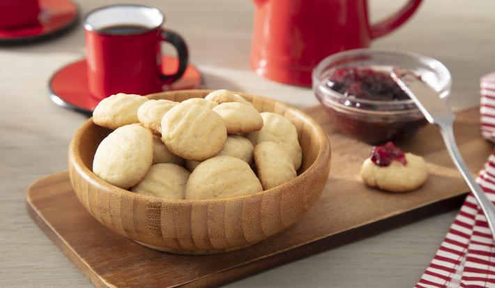 Foto da receita de biscoito de nata. Observa-se os biscoitinhos dentro de um recipiente fundo e redondo de madeira. Ao lado, um dos biscoitinhos está com geléia de morango e atrás tem uma xícara com café.