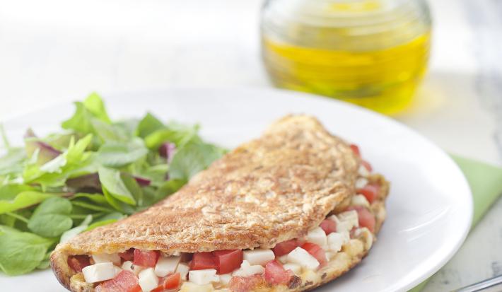 Fotografia em tons de verde em uma mesa de madeira cinza com um pano verde, um prato branco raso redondo e a omelete integral recheada com queijo branco, tomate e ervas, ao lado salada para acompanhar. Ao fundo, um potinho com azeite.